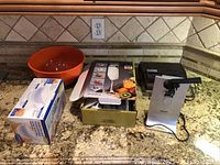 Overview photo showing all lot items on a kitchen counter including the Krups electric can opener on the right, Snakmaster sandwich grill next to it, boxed two-speed hand mixer in the middle, and metal red popcorn bowl on the left.