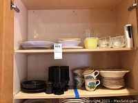 View of upper and lower shelves containing stacked plates, bowls, glass candle holders, cups, and small containers in various materials and colors.