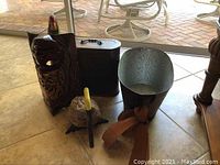 Five pieces of room decor on tile floor near patio door: metal bucket with ribbon, carved wooden mask, black metal bell, and carved pelican and parrot sculptures.