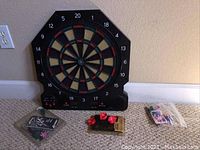 Black octagonal electronic dartboard with digital scoreboard on bottom and darts and flights in packaging laid out on carpet.