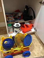 View inside a cabinet shelf showing an assortment of dog accessories including a ceramic treat container, orange harness, brush, water bottle, and a yellow plastic wagon filled with toys.