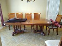 Wide view of the rectangular wood dining table with six chairs, showing the entire tabletop, pedestals, and included custom table pads stacked on one end.