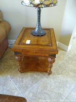 Wood side table shown with a lamp base on top (lamp not included). Shows top inset panel, carved legs, and lower shelf.