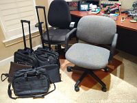Two office chairs, gray and black, with armrests and wheels, placed side by side along with multiple black briefcases and roller carry-on luggage. Clear floor protector mat is beneath the chairs.
