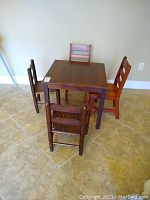 Overview of square wooden child table surrounded by four wooden chairs with ladder backs, dark brown finish showing wear