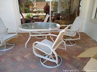 Four white metal rocking patio chairs arranged around a glass top patio table on a brick patio.