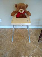 Full view of cream plastic high chair with metal legs and large brown teddy bear seated in it on tiled floor.