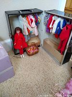 Wide shot of the open black Barbie trunk with doll clothing hanging inside, Skipper doll in red coat to the side, small basket of doll hangers, and accessories box on carpet floor.