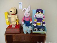 Wide view of Snoopy phone, rapping bunny doll in box, and small light blue plush bunny on wooden surface