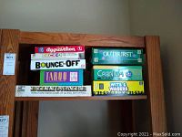 Top shelf of wooden bookshelf holding stacked adult board games including Aggravation, Bounce-Off, Taboo, Billionaire and more.