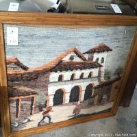 Large framed woven wall art showing a scene of a mission or hacienda with people walking in front, framed in medium oak.