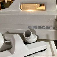 Close-up of Oreck XL hand vacuum with visible attachments including brush and nozzle; vacuum casing is light gray with visible Oreck XL branding