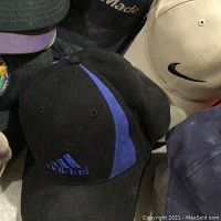 Photo showing black Adidas cap with blue stripe and logo, beige Nike cap with black swoosh, and more caps behind.