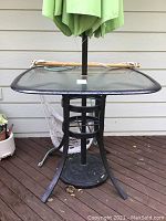 Photo of black metal patio table with glass top and folded green umbrella attached in the center.