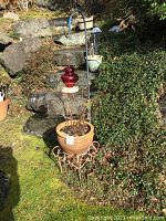 Large terra cotta planter with Greek key design on a metal stand, containing soil and a plant, with a shepherd's hook holding a red glass hummingbird feeder.