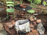 View of round black metal bistro table with decorative floral edge patterns, two green metal chairs with a simple ladder-back design, shallow terra cotta planter on table, and small garden cart with fabric bag and handle in outdoor setting.