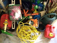 Photo showing an assortment of garden supplies including a red gas can, yellow extension cord, gardening book, tools, gloves, and a green watering can with flexible spout.
