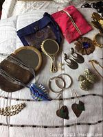 Photo showing assorted jewelry pieces laid out including fabric pouches, earrings, necklaces, pins, bracelets, and a small mirror.