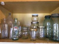 Display of assorted clear and green glass canning jars and vintage bottles on wooden shelf.