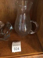 Clear glass Princess House pitcher with flared top and handle, displayed in wooden shelf.