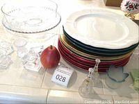 Photo showing assorted charger plates stacked including white, green, and red colors, clear glass napkin rings, clear decorative glass pitchers, and other glassware on a counter.