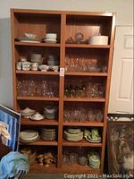 Front view of double wide light oak bookcase filled with assorted glassware and dishware.