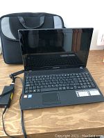 Open Emachines E443-BZ844 laptop showing keyboard, screen, power cord plugged in, and black padded carrying case behind it.