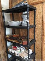 Two black plastic utility shelving units placed in garage-like space, one with various miscellaneous items on shelves (not included).