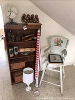 Overview of bookshelf filled with various items including wicker baskets, lamp base, hangers, beach umbrella, and vintage wooden high chair with bear painting.