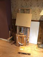 Large wooden standing easel along with wooden folding easel and case, two canvases leaning against the wall.