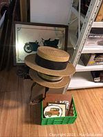 Vintage wicker hats stacked on top with framed vintage car plaque in background, along with other memorabilia items in a green crate nearby.