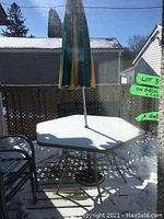 White metal patio table with umbrella and umbrella base visible on a wooden deck with snow and fence background.