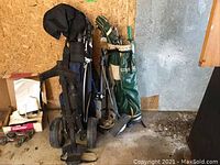 Two golf bags and two pull carts against a plywood and concrete wall, left the blue bag and right the green bag. Two umbrellas visible attached to bags.