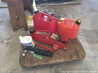 Photo showing red hydraulic floor jack, two red gas cans, black trailer hitch ball mount, and white bottle on wooden surface.