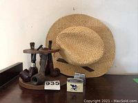 Five smoking pipes on a wooden two-tier stand with a wicker hat behind it and tag number 935