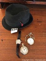 Overview of all four items on a wooden surface; includes black felt hat, two pocket watches, and wristwatch.