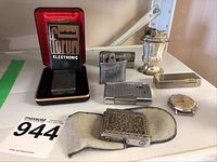 Group photo showing six vintage lighters and one wrist watch on white surface including Forum Electronic lighter in its fitted case, ornate lighter with pouch, urn-shaped lighter, and Timex wristwatch without band.