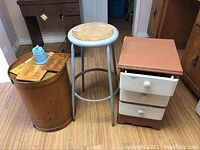 Front view of cheese barrel, metal stool, and three-drawer side table arranged next to each other on wood floor