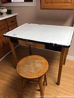 Full view of metal utility table with wooden legs and single drawer paired with round wooden stool showing surface wear.