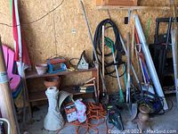 Wide view showing wooden shelving unit, various gardening tools, hoses, pots and extension cords stored against a wall