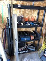 Black plastic utility shelf with three shelves against wooden wall containing assorted hand tools, cases, and measuring and leveling tools.