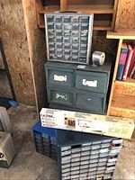 Photo showing two stacked metal filing boxes with wear and three plastic hardware caddies containing hardware, plus boxed fluorescent light fixture on floor next to filing boxes.