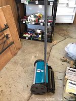 Full view of blue push reel lawn mower with handle extended on concrete floor near shelving.