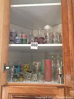 Glass cabinet showing multiple shelves of assorted retro glassware items including shot glasses, decorative drinking glasses, and a pitcher.