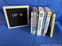 Front view of seven DVD cases of The Office series 1 through 5 and two extra DVDs placed upright next to a black felt board with white letters reading LOT 16.
