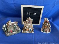 Photo of three porcelain Christmas village log cabins arranged on blue cloth background with letter board reading 'Lot 19' behind