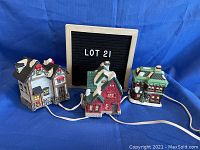 Three porcelain Christmas village buildings connected with electrical cords for lights. Includes a white building with black roof and chimney, a red building with snow-covered green roof, and a green building with wreath and snow accents.