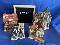 Four Christmas village buildings displayed on blue fabric background: General Store with red roof, church with stone facade, coffee shop with green roof, and cottage with brown/yellow roof.