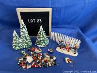 Image showing four ceramic Christmas trees, a white picket fence, a figurine group of three children on a snow base and numerous hanging ornaments scattered in front.