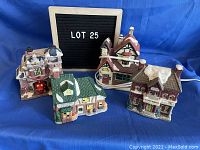 Photo of four ceramic Christmas village buildings on blue backdrop, showing Fire Station, Grand Hotel, Village Hospital and Grocery Store.